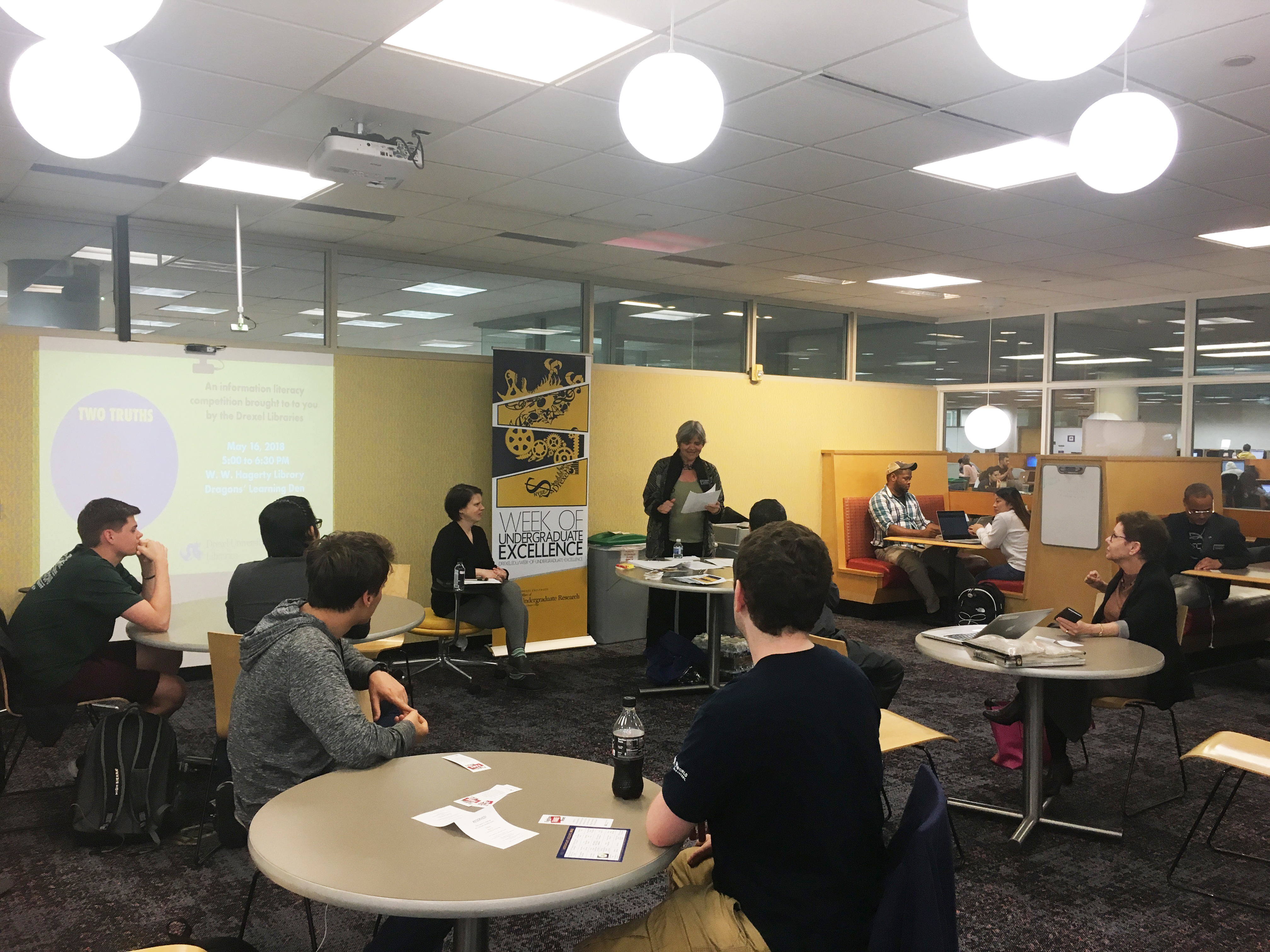 Students sit at round tables and face a woman standing at the front of the room.